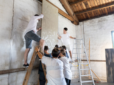 Das Team bringt mit vereinten Kräften über Leitern eine strukturierte Leinwand an der Wand der alten Fabrik an